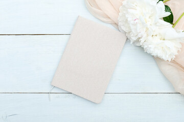 Paper notebook and beige peony flowers on blue wooden table top view. Feminine workspace, flat lay style composition.