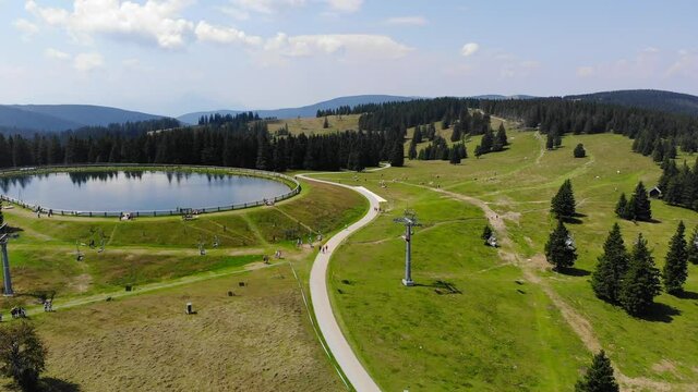 Mountain sports resort in Rogla Slovenia with ski lift chairs and round lake with people enjoying a sunny day, Aerial flyover shot