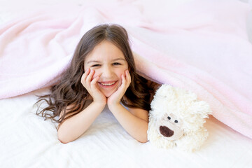 a small smiling girl 5-6 years old is lying in bed with a Teddy bear, hands under her cheeks