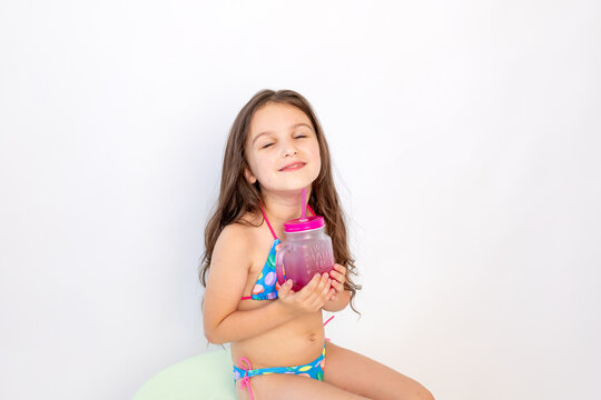 a little girl of 5-6 years old is sitting in a swimsuit with a cocktail on a white isolated background, a place for text