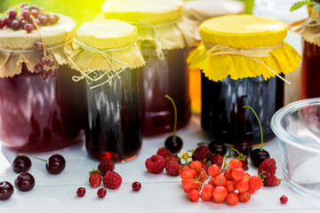 Fruit and berry preserves in the glass jars and raw strawberries , cherries , rowans and red currants berries on a white wooden table, on the nature background, organic meal and dessert concept