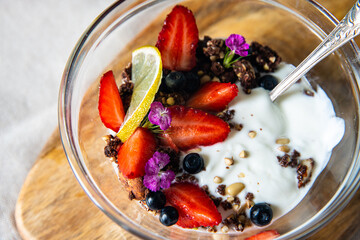 Yogurt with granola, bilberries, strawberries, seeds, pine nuts and sweet William flowers in glass bowl on wooden board on white background.  