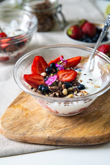 Yogurt with granola, bilberries, strawberries, seeds, pine nuts and sweet William flowers in glass bowl on wooden board on white background.  
