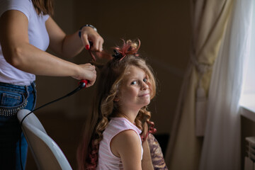 Woman making a hairdressing by curly iron to young cute girl with long hair indoor. Curly style.
