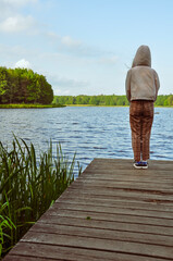 Samotna dziewczyna na spacerze stojąca tyłem w kapturze i spodniach w kratę nad brzegiem jeziora. 