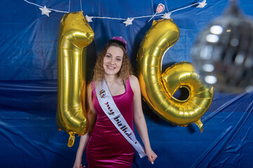 Sixteen year old girl wearing a tiara and sash celebrates her birthday with a party featuring gold...