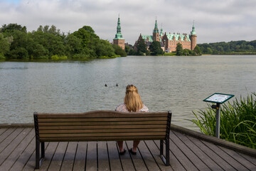 Fototapeta na wymiar Blond girl sitting on a bench and looking at a beautiful Frederiksborg Castle and a lake around it, in Hillerød, Denmark
