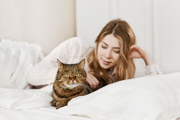 A charming smiling girl strokes her furry pet cat while lying on the bed in the morning. Pets, care, care. Side view, space for text