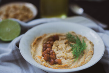 Chickpea vegan hummus garnished with dill and baked chickpeas on black background with vintage spoon, bottle of olive oil, towel, chickpeas and half a lime