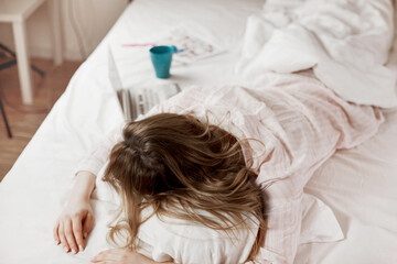 A young attractive girl in pajamas is lying on the bed at home, next to a laptop and a Cup of coffee. The girl works before going to bed. The view from the top