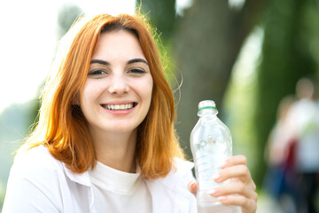 Thirsty smiling redhead woman drinking water from a bottle in summer park.