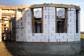 House under construction, walls of white foam aerated concrete blocks, wooden scaffolding, big window openings and concrete foundation.
