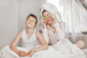 Obraz na płótnie Canvas A beautiful young girl and her cheerful brother make a face mask from cucumber slices. The brother and sister are sitting on the bed with cucumbers in their eyes. The concept of beauty and health