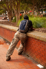 an African American backpacker  in camouflage pants is leaning against a brick wall in an urban are