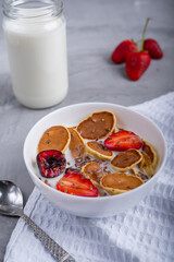 White bowl full of milk with mini pancakes and berry mix laying on white napkin near silver spoon and jar with milk on concrete surface at kitchen