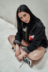 Young and cute female model posing on the white background while sitting on the floor and wearing casual clothes with custom sneakers, full height portrait