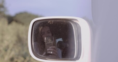 Young adult female taking selfie in rear view mirror of Camper van on road trip at sunset