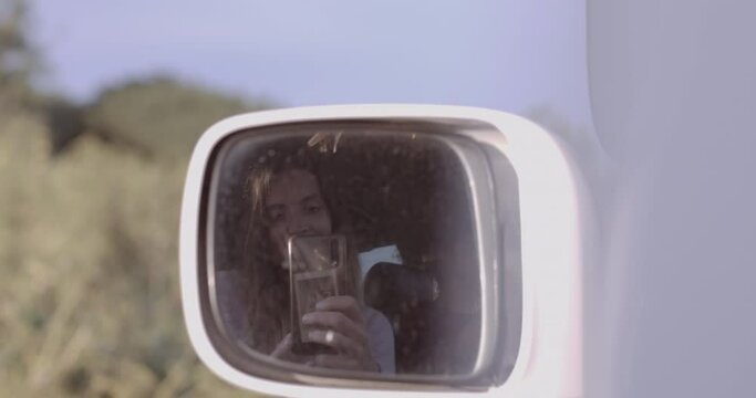 Young adult female taking selfie in rear view mirror of Camper van on road trip at sunset