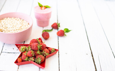 Fresh strawberry yogurt with berries around on a white wooden background. A pink shake in a glass, next to it is a pink cup with Hercules flakes, and red strawberries lie in a red star-shaped plate