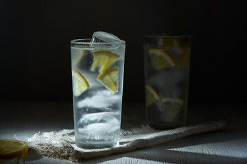 Two glasses of ice cold lemonade with lemon slices and ice on linen napkin on black background in blur.