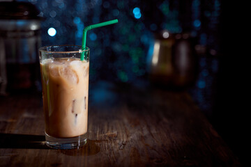 Ice coffee in a tall glass with cream poured over, ice cubes and beans on a old rustic wooden table. Cold drink with tubes on a black background