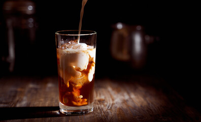 Ice coffee in a tall glass with cream poured over, ice cubes and beans on a old rustic wooden table. Cold drink with tubes on a black background