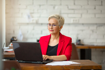 beautiful aged woman in a red jacket works in the office with a laptop