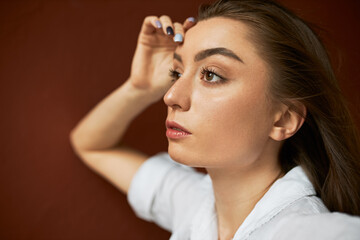 Naklejka premium Close up image of attractive thoughtful young Caucasian woman with clean skin and loose brown hair touching face, having pensive facial expression, looking ahead of her, recollecting something