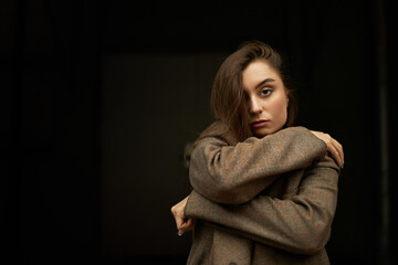 Attractive confident young European woman staring at camera with serious look keeping arms around shoulders, hugging herself, wearing oversize jacket, posing against black copy space background
