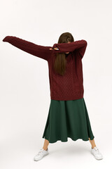 Full length vertical image of unrecognizable young dark haired woman wearing white sneakers, green skirt and knitted sweater posing isolated making dancing gesture, covering face with ellbow