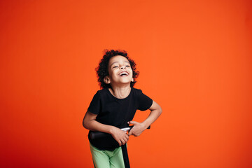 Joyfully laughing boy in black t-shirt riding on scooter on his hands and head on an orange background in studio