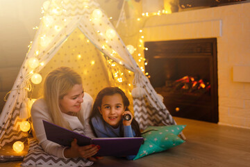 Obraz na płótnie Canvas Mother and daughter are sitting in a teepee tent, reading stories with the flashlight. Happy family.