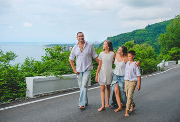 Fototapeta premium Cheerful, happy family, father, mother, son and daughter enjoying walk play outdoor near sea. Family in white clothes barefoot on walkway. Green forest background.