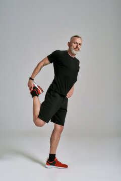 Full Length Shot Of Middle Aged Muscular Man In Black Sportswear Looking At Camera While Stretching His Body Before Workout In Studio Over Grey Background