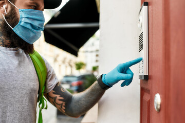 Brutal bearded delivery man wearing mask and gloves due to the emergence of the Covid19 virus, ringing a doorbell, while delivering food. Courier, delivery service, lockdown concept
