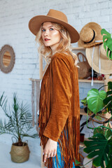 Fashionable woman wearing boho outfit: wide bream hat, brown suede jacket with fringe, posing in interior with trendy decor © Victoria Fox