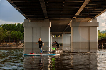 Fototapeta na wymiar View on man and woman on sup boards under bridge.
