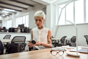 Beautiful woman at work. Portrait of young and cute blonde tattooed business woman adjusting her makeup, looking in the small mirror while sitting in the modern office