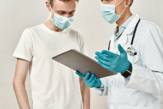 Doctor And Young Patient In Medical Masks. Mature Male Doctor In Medical Uniform Holding Digital Tablet, Explaining Diagnosis To A Young Man