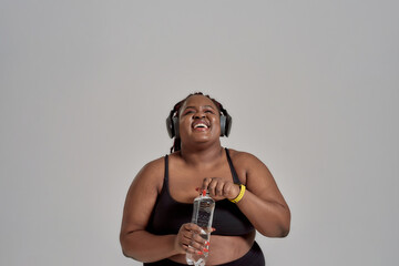 Tomorrow is looking great. Plump, plus size african american woman in sportswear and headphones laughing, opening bottle of water, posing in studio over grey background
