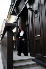 Outdoor photo of brunette lady walking on street background in cold autumn day. Fashion street style portrait. wearing dark casual trousers, white shirt, sunglasses and black coat. Fashion concept.