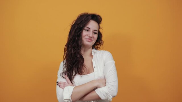 Standing and cross her arms on chest. Young attractive woman, dressed white blouse, with brown eyes, curly hair, yellow background