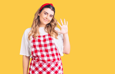 Young caucasian woman wearing apron showing and pointing up with fingers number five while smiling confident and happy.