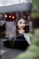 Obraz na płótnie Canvas Beautiful brunette girl behind the glass, portrait of young woman in window, concept of female emotions. female model looking through the window. Winter cafe concept.
