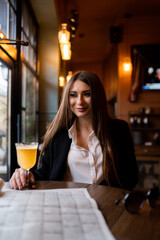Beautiful woman with brunette hair in dark clothes looking at window and drinking orange hot drink. Concept of cozy places and cold winter. Concept of cafe and coffee shop. Cosy and atmosphere place.