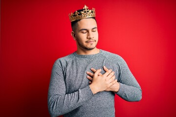Young handsome man wearing golden crown of prince over isolated red background smiling with hands...