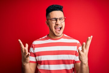 Young handsome man wearing casual striped t-shirt and glasses over isolated red background celebrating mad and crazy for success with arms raised and closed eyes screaming excited. Winner concept