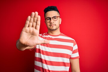 Young handsome man wearing casual striped t-shirt and glasses over isolated red background doing stop sing with palm of the hand. Warning expression with negative and serious gesture on the face.