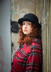 Portrait of a young woman with curly red hair, wearing a red tartan coat, set in a rough urban setting.