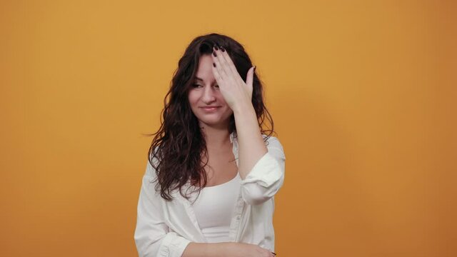 Closing face palm covers eyes visible through with hands. Young attractive woman, dressed white blouse, with brown eyes, curly hair, yellow background